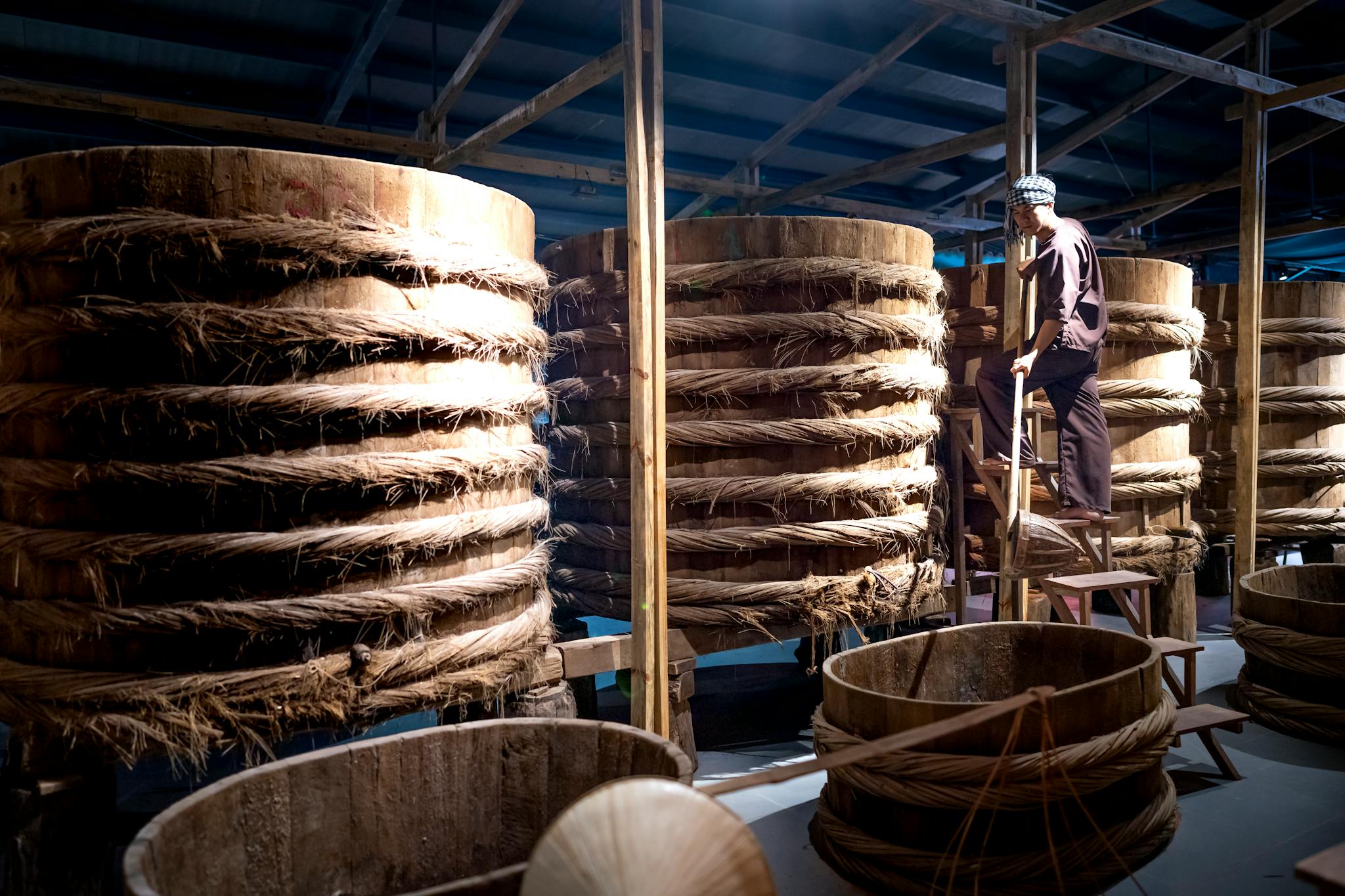 A worker oversees large wooden barrels used for traditional fermentation in an indoor warehouse setting.