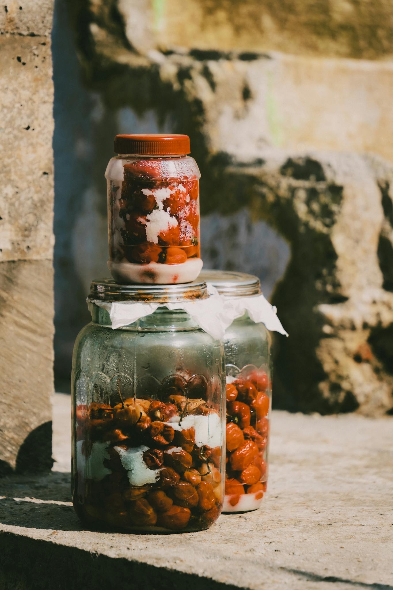 Close-up of glass jars with fermented fruits and sugar on rustic stone surface outdoors.
