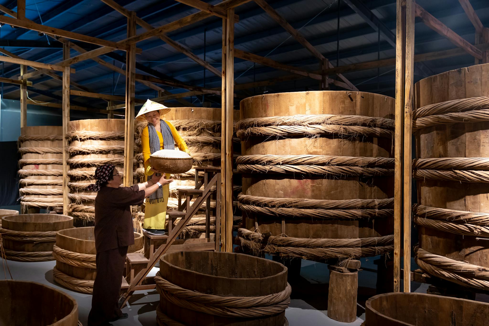 Workers in a traditional Asian fermentation facility managing wooden barrels for food production.