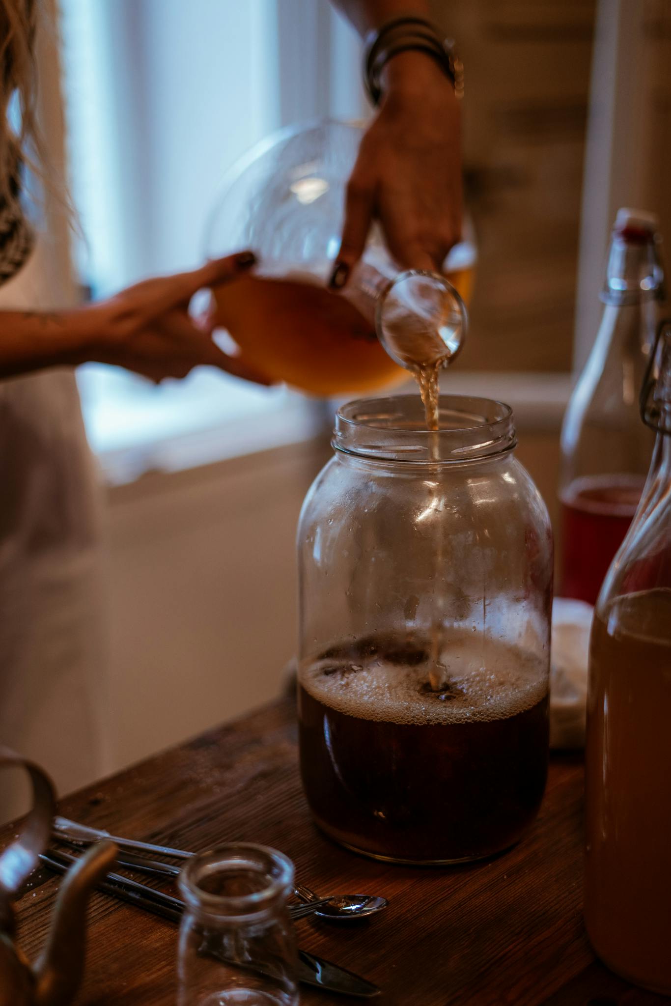 A person pouring homemade kombucha into a glass jar on a wooden table indoors.